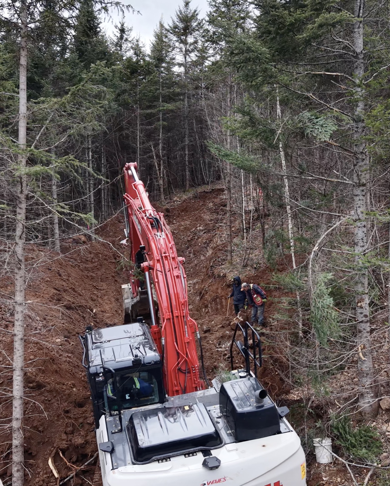 Excavator cutting an access trench through forest while two crew members inspect the wall